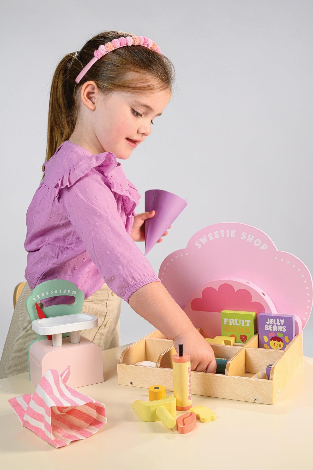 A young girl plays with the Tender Leaf Wooden Toys Sweetie PicnMix Shop, featuring colorful compartments filled with treats. The shops pastel design enhances its charming, retro aesthetic.