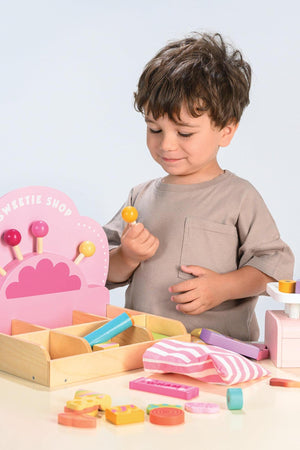 A child in a beige shirt interacts with a colorful wooden Sweetie Shop toy, featuring pink and yellow components.