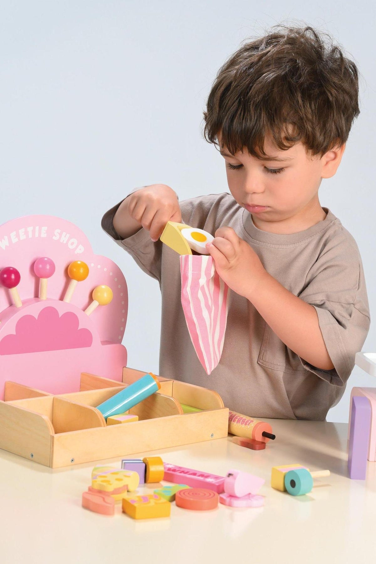 A young child is playing with the Tender Leaf Wooden Toys Sweetie PicnMix Shop, using a colorful scoop to select treats from a wooden compartmentalized set on a light-colored table.