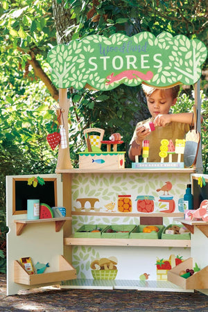 Child playing with Tender Leaf Woodland Stores playset, featuring colorful wooden fruits and a green forest backdrop.