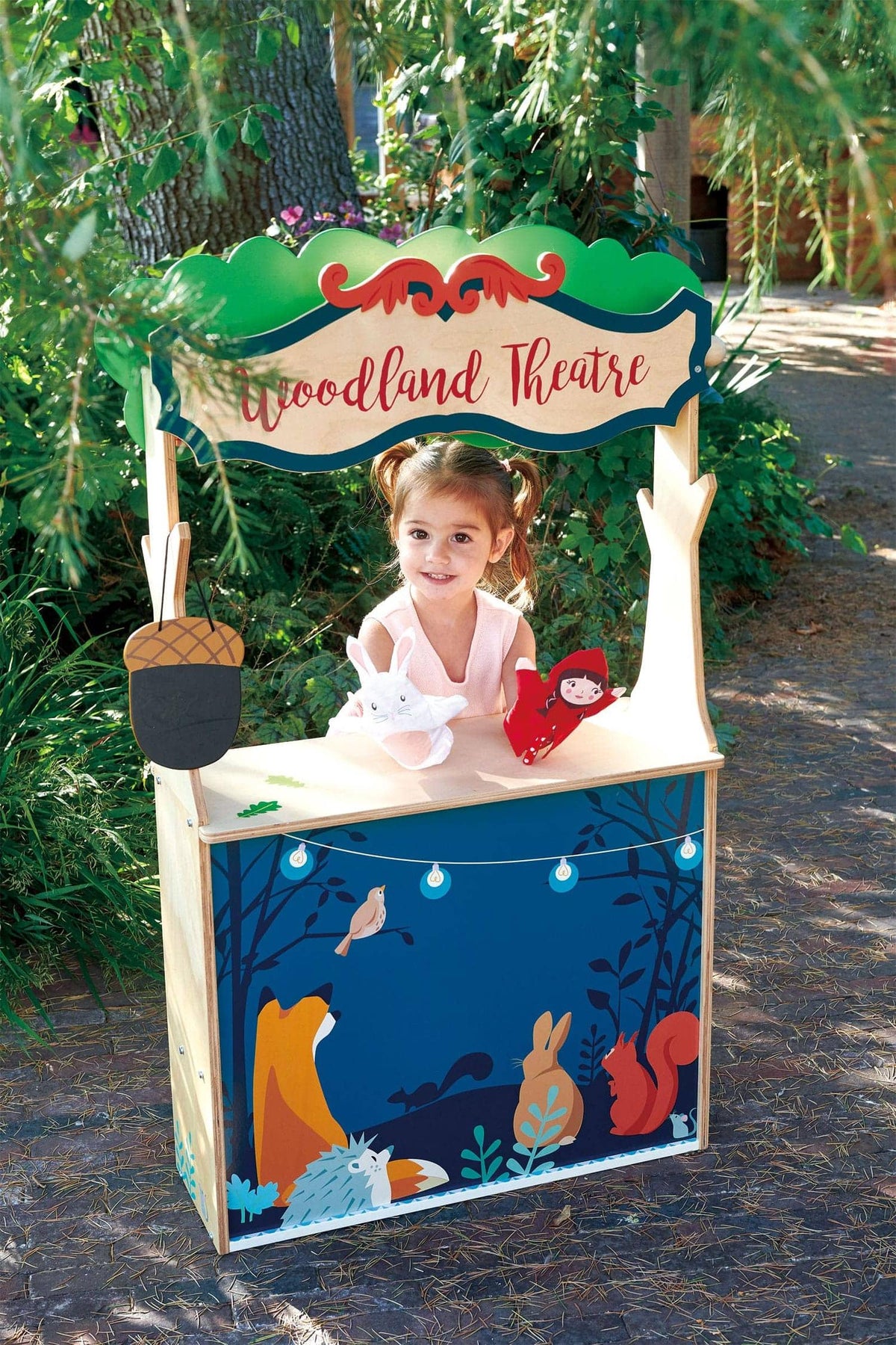Child playing at a colorful wooden Woodland Theatre playset outdoors, with animal illustrations and puppet toys.