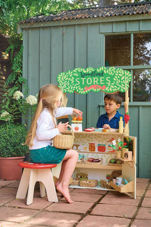 Two children play with a Tender Leaf Woodland Stores wooden toy set outdoors, featuring shelves and a colorful sign.