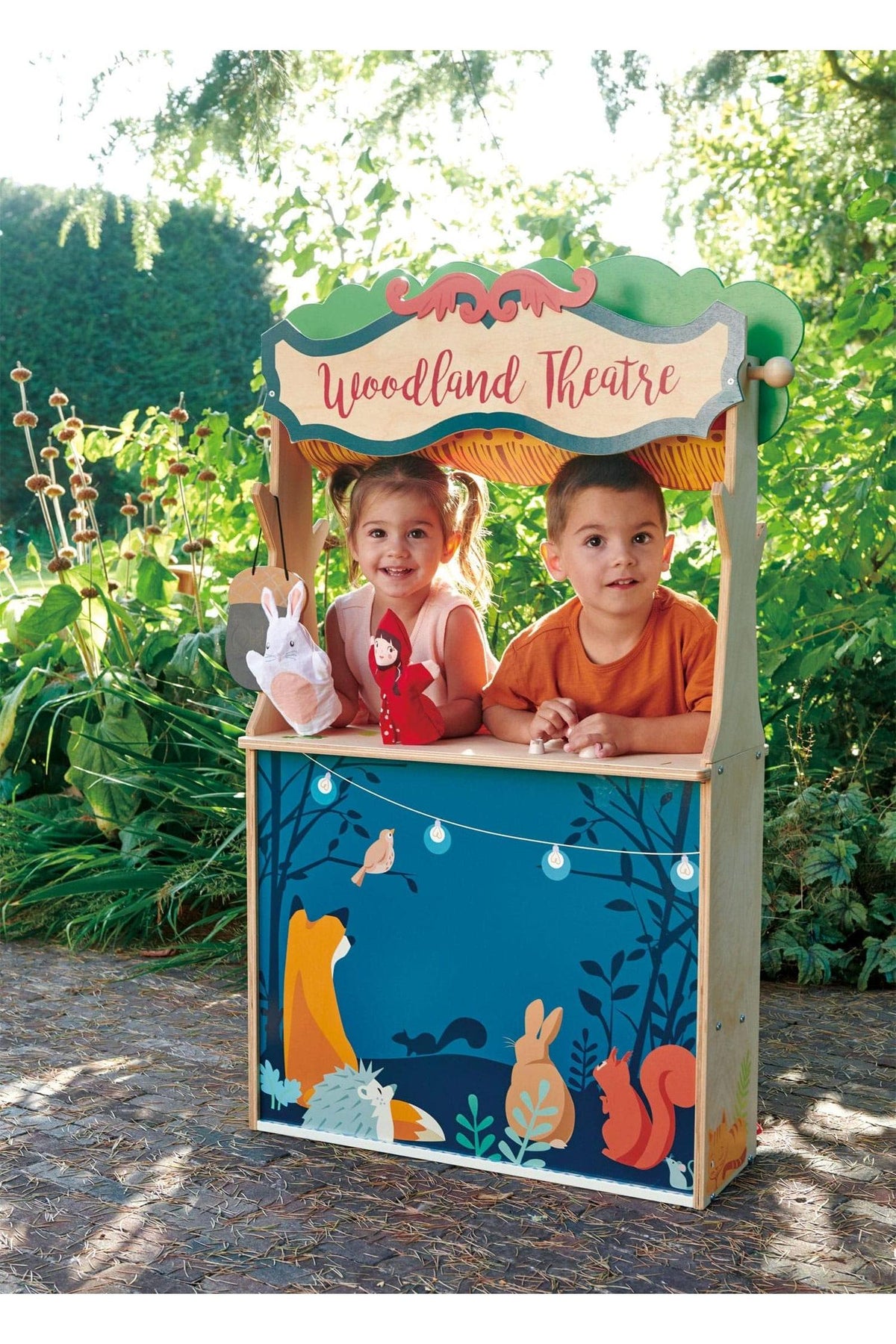 Two children play at a colorful wooden Woodland Theatre with forest animal illustrations, outdoors in a garden setting.