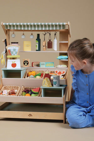 A young child interacts with a wooden market stall filled with colorful play food, showcasing sustainable materials and imaginative play.