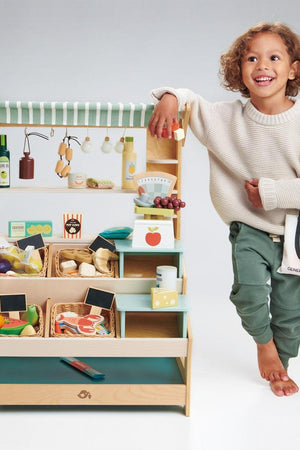 A child stands next to a colorful wooden market stall filled with various toy fruits, vegetables, and accessories.