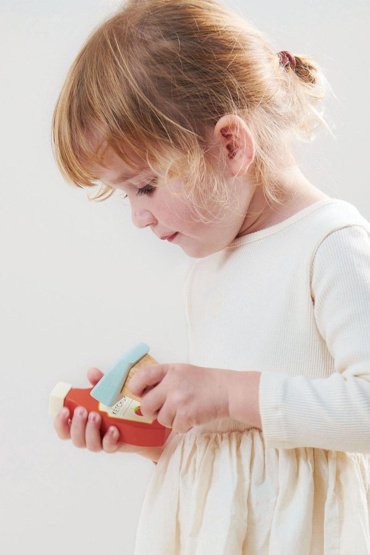 A child in a cream dress plays with a colorful wooden toy scanner, focused and smiling.