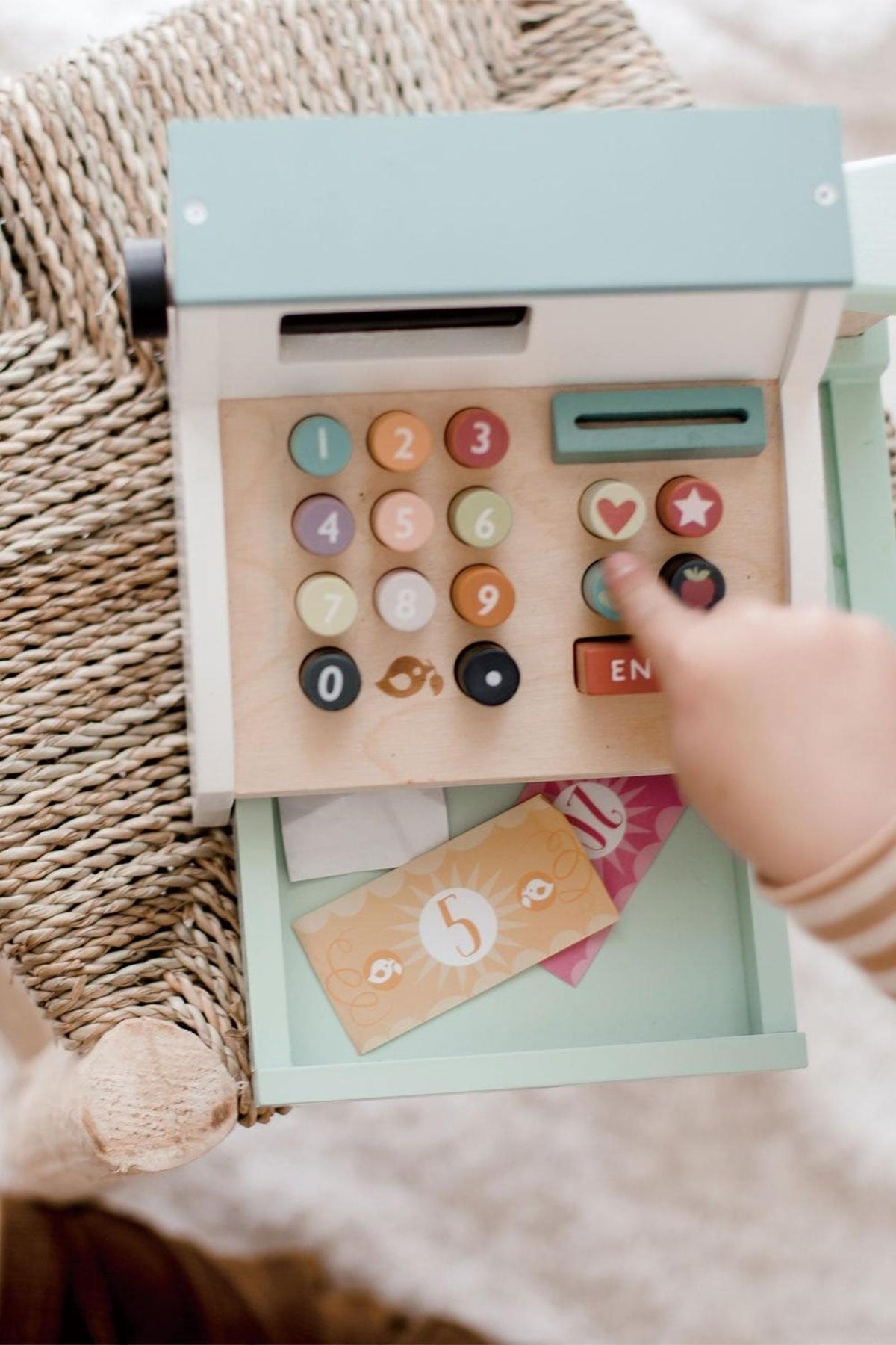 A child interacts with a colorful wooden cash register toy on a woven surface, featuring buttons and play money.