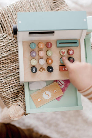 A child interacts with a colorful wooden cash register toy on a woven surface, featuring buttons and play money.