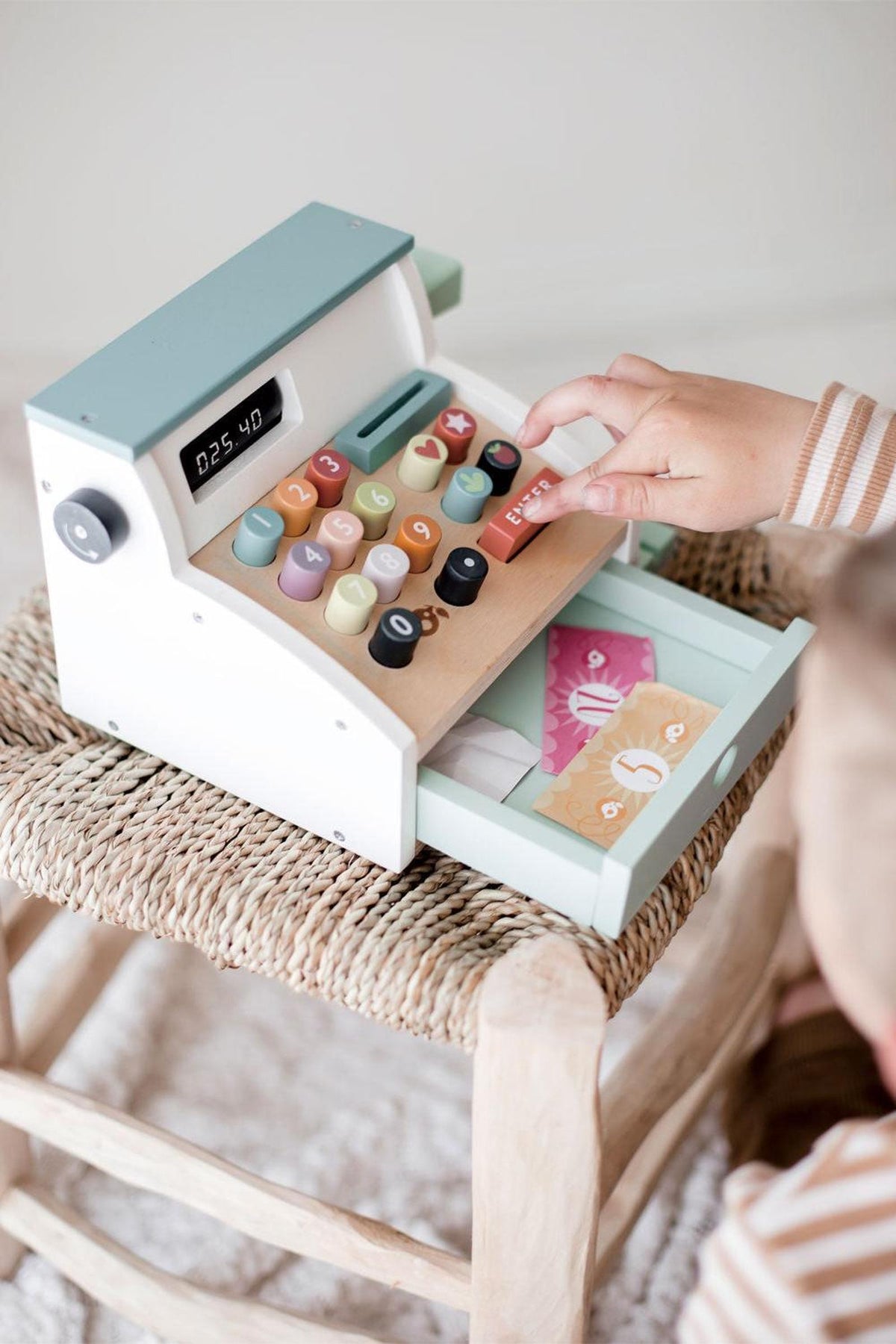 A child interacts with a colorful wooden cash register on a woven stool, featuring buttons and a drawer.