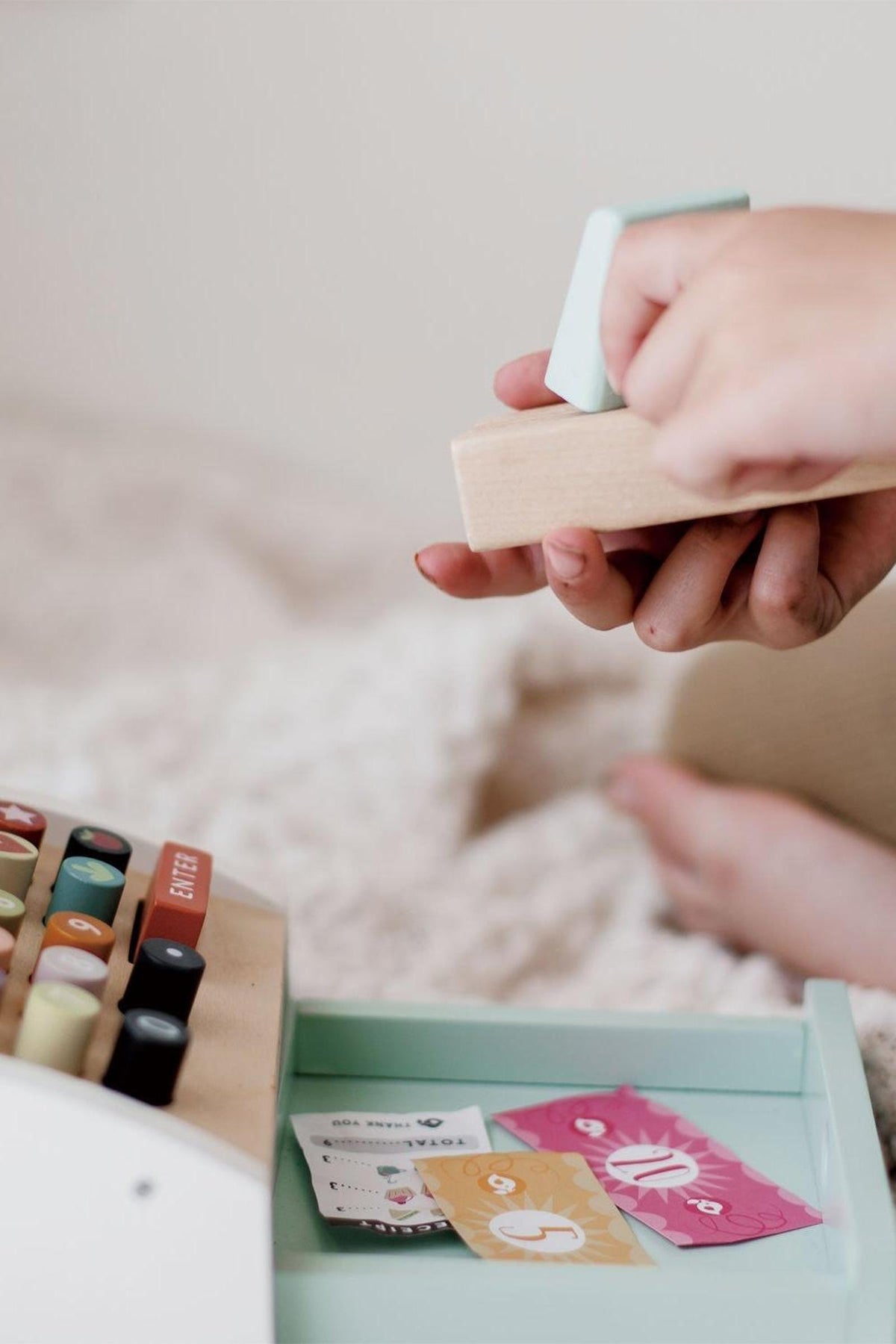Child playing with a wooden cash register, featuring colorful buttons and paper money, on a soft surface.