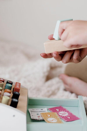 Child playing with a wooden cash register, featuring colorful buttons and paper money, on a soft surface.