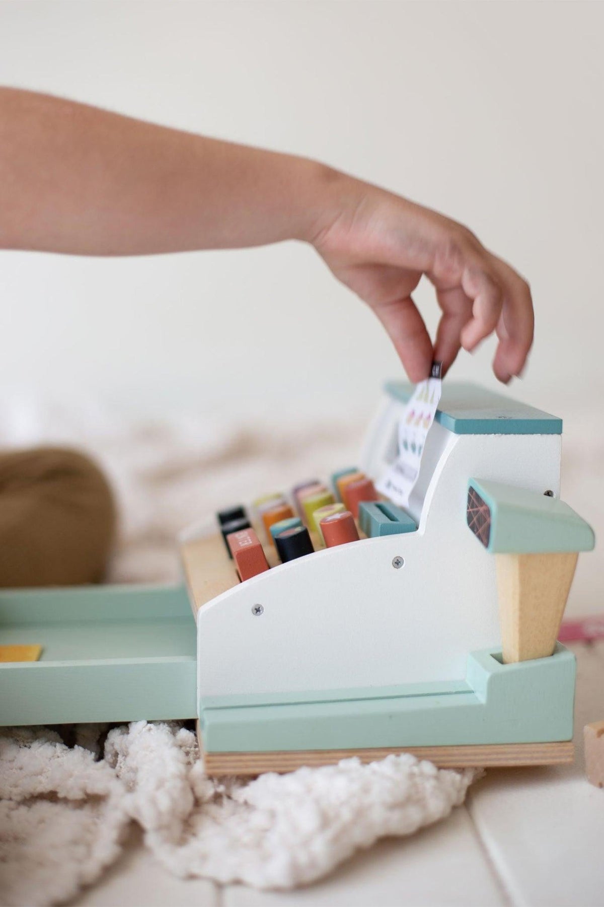 A child interacts with a pastel-colored wooden cash register toy, featuring colorful buttons and a small receipt.