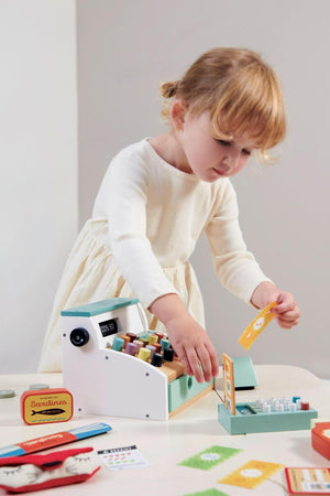A child plays with a colorful wooden cash register, featuring buttons and accessories, on a light-colored table.