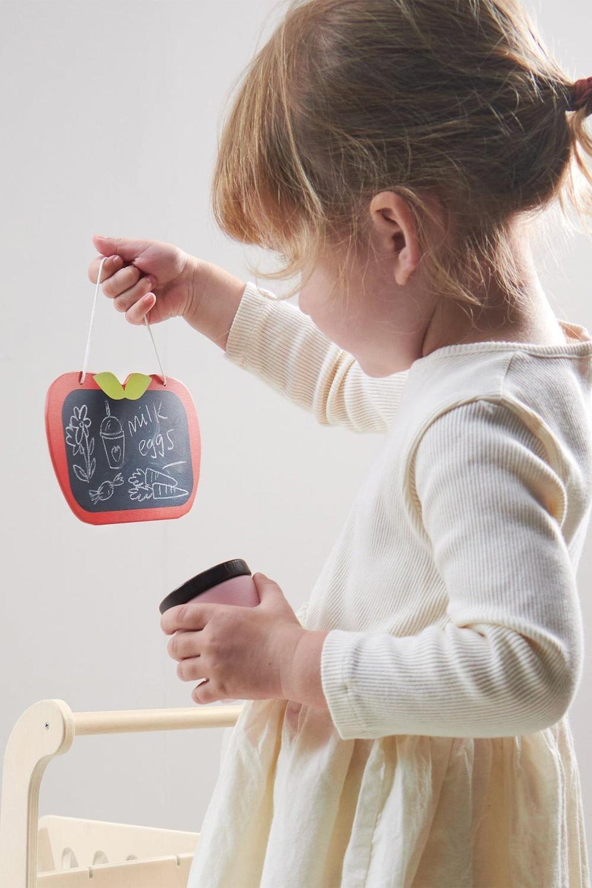 A young child holds a red apple-shaped chalkboard with drawings, showcasing playful creativity and engagement in pretend shopping.