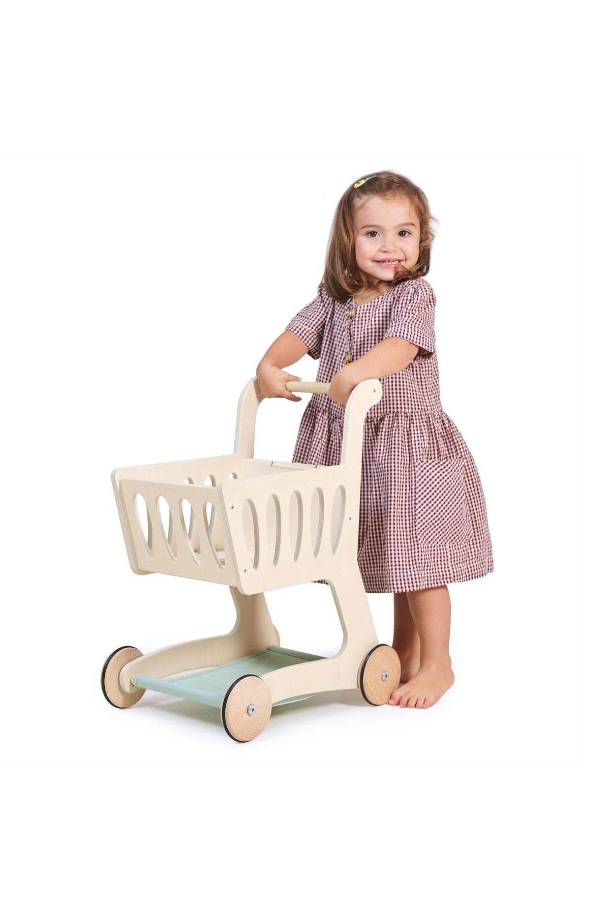 A young girl in a checkered dress stands barefoot, pushing a light-colored wooden shopping cart with a spacious basket.