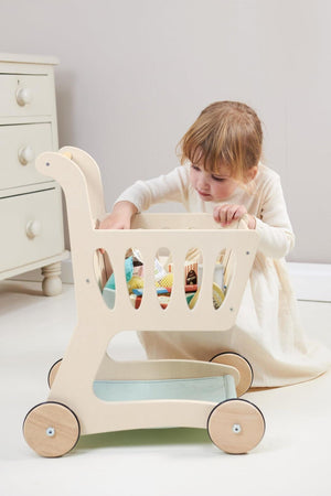 A young child in a cream dress plays with a wooden shopping cart, filled with colorful toys, on a light.