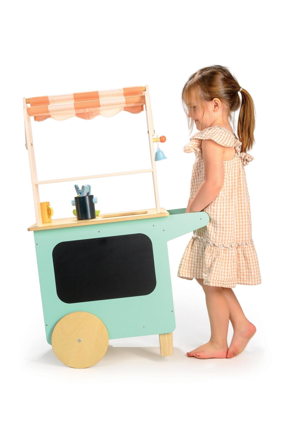 A child plays with a Tender Leaf Wooden Toys drinks trolley, featuring a striped fabric awning and wooden wheels.