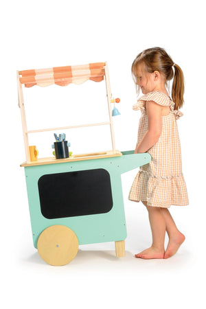 A child plays with a Tender Leaf Wooden Toys drinks trolley, featuring a striped fabric awning and wooden wheels.
