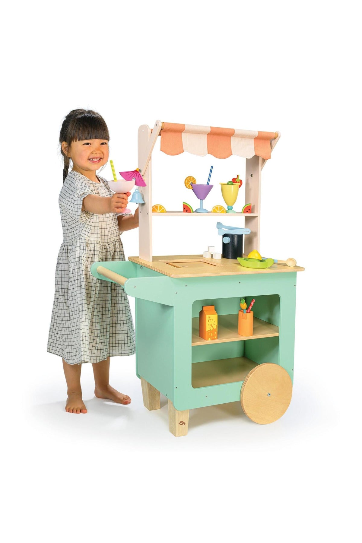 A child plays with a Tender Leaf Wooden Toys drinks trolley, featuring colorful mocktail accessories and a striped awning.