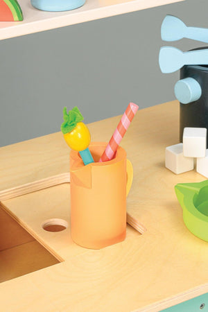 Wooden orange cup with a pink straw and fruit stirrer on a light wooden surface, alongside colorful accessories.