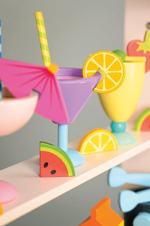 Colorful wooden drink set with purple and yellow glasses, fruit slices, and straws on a white shelf.