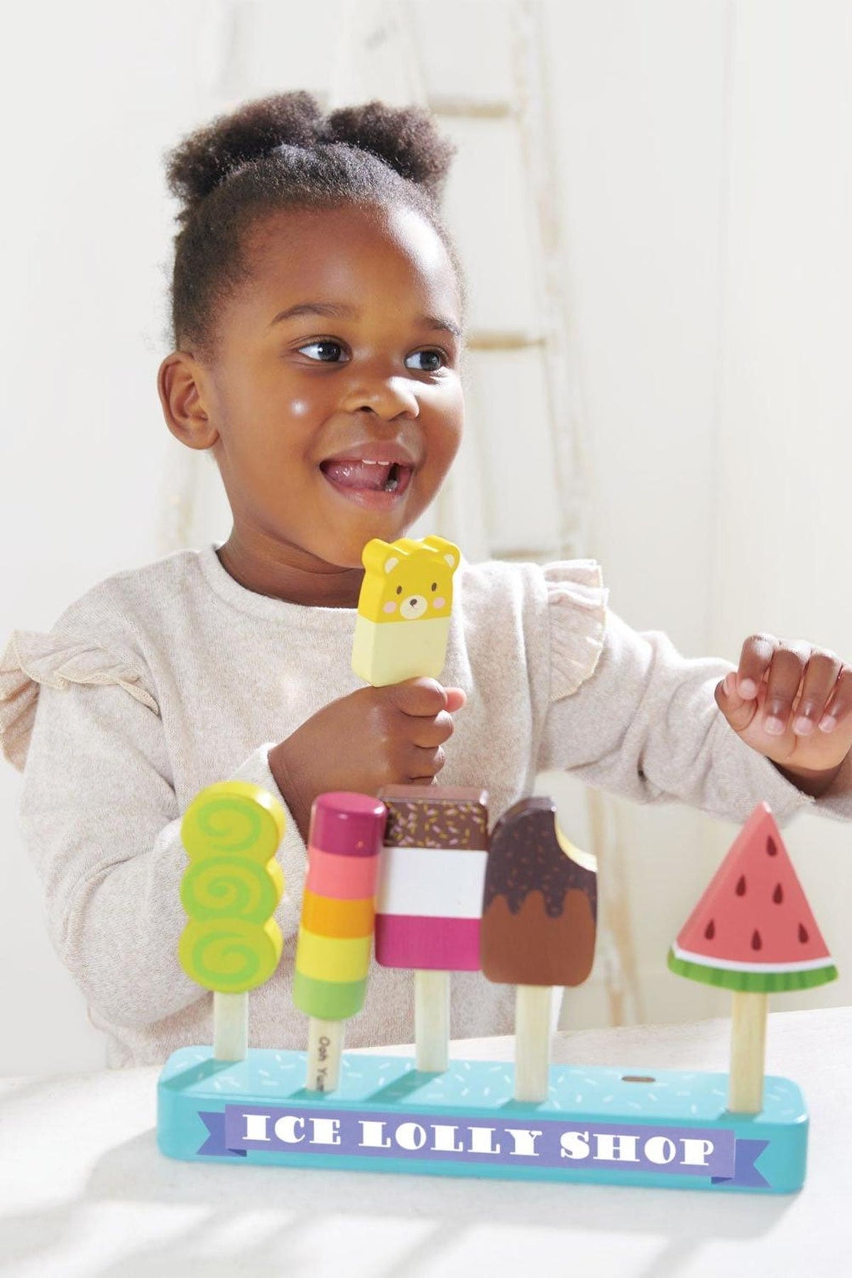 A smiling child holds a popsicle in front of a colorful wooden ice lolly shop displaying various toy popsicles.