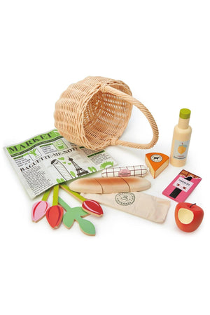 A wicker shopping basket contains wooden toys, including tulips, a baguette, cheese, a lemonade bottle, and a newspaper.