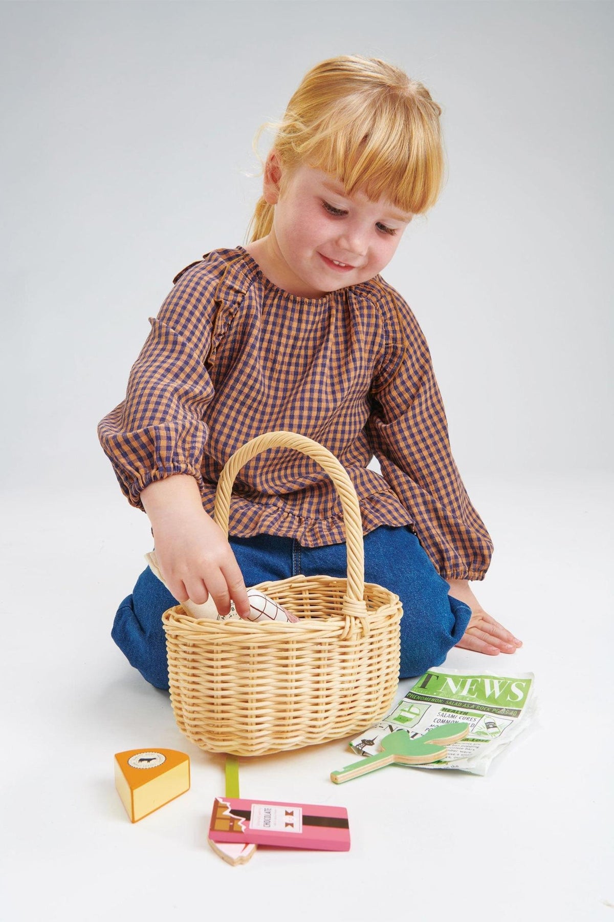 A young girl in a checkered blouse sits on the floor, reaching into a wicker basket filled with colorful toy.