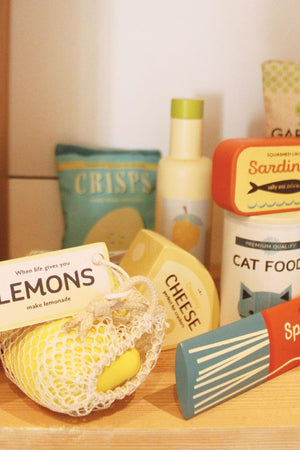 Colorful wooden grocery items are displayed on a shelf, including cheese, cat food, and a string bag with lemons.