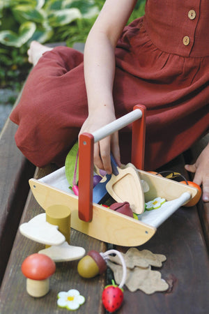 A child in a rust-colored dress plays with a wooden foraging trug filled with colorful, eco-friendly wooden pieces and leaves.