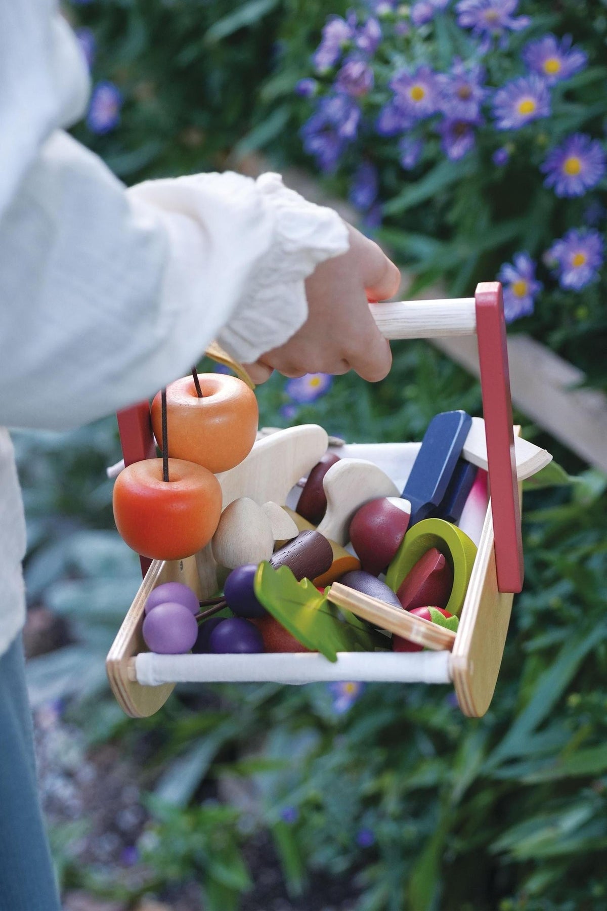 A child holds a wooden trug filled with colorful, eco-friendly toy fruits and vegetables, surrounded by vibrant greenery and flowers.