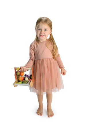 A young girl in a pink dress holds a wooden trug filled with colorful toy fruits and vegetables.