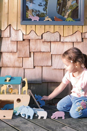 A young girl interacts with colorful wooden animal figures near a large wooden ark, set against a rustic wooden backdrop.