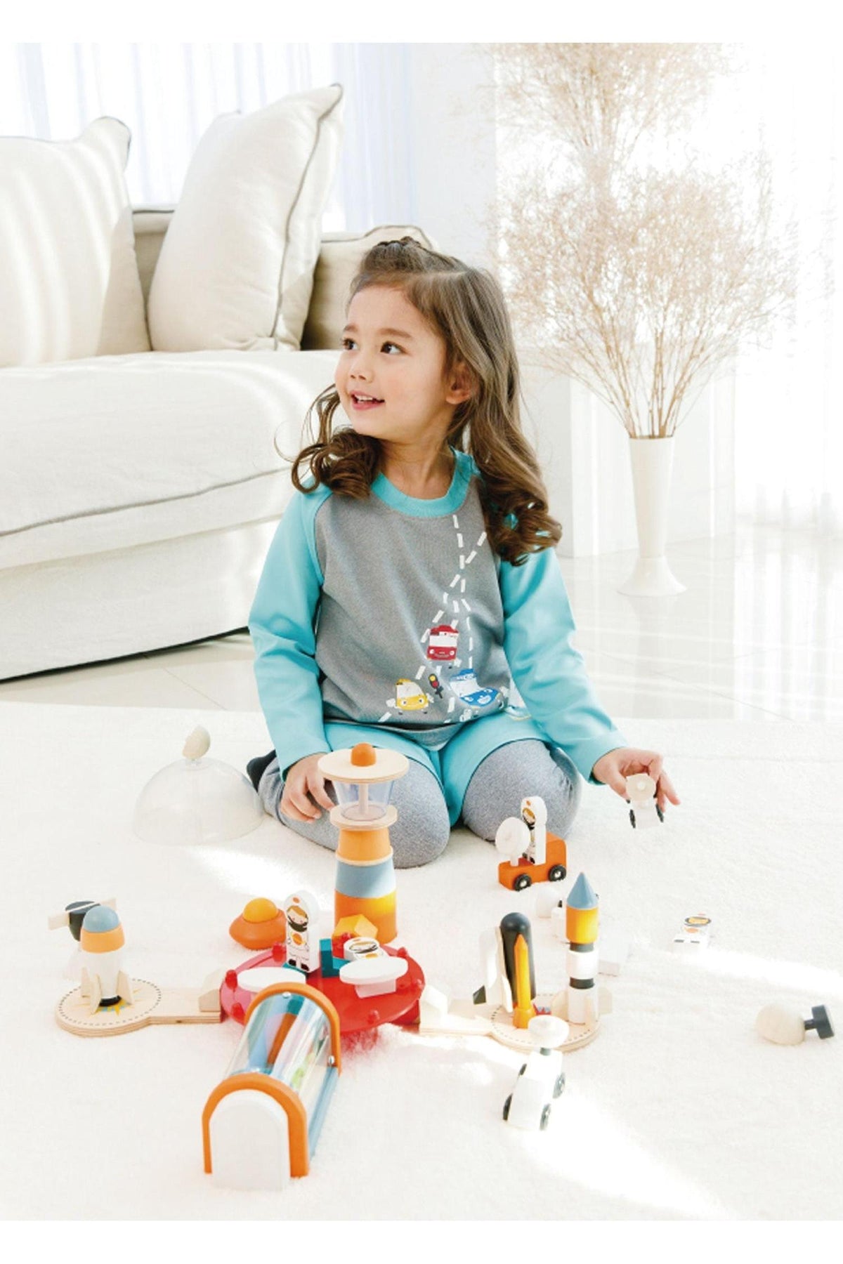 A young girl smiles while playing with a colorful wooden toy set on a light-colored carpet in a bright room.