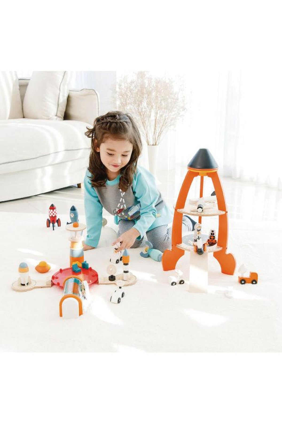 A young girl plays with a colorful wooden rocket set on a bright, airy floor, surrounded by imaginative space-themed figures.