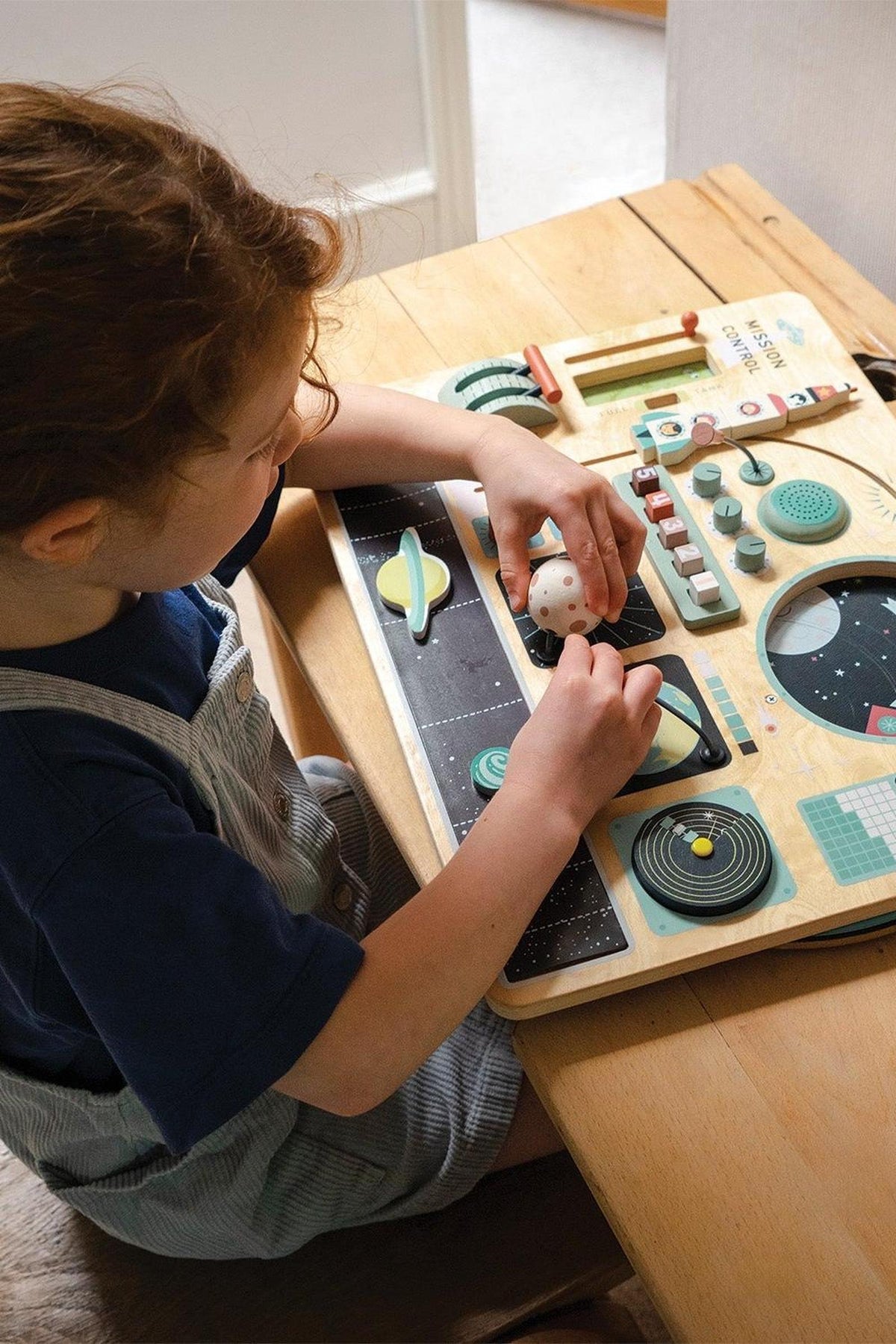 A child interacts with a wooden space station toy on a table, featuring colorful dashboard activities and rotating elements.