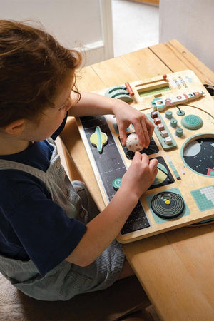 A child interacts with a wooden space station toy on a table, featuring colorful dashboard activities and rotating elements.