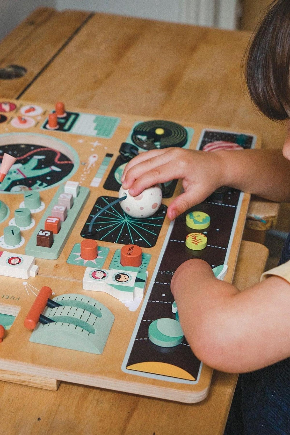A child interacts with a colorful wooden space station toy, featuring various buttons and activities on a plywood board.