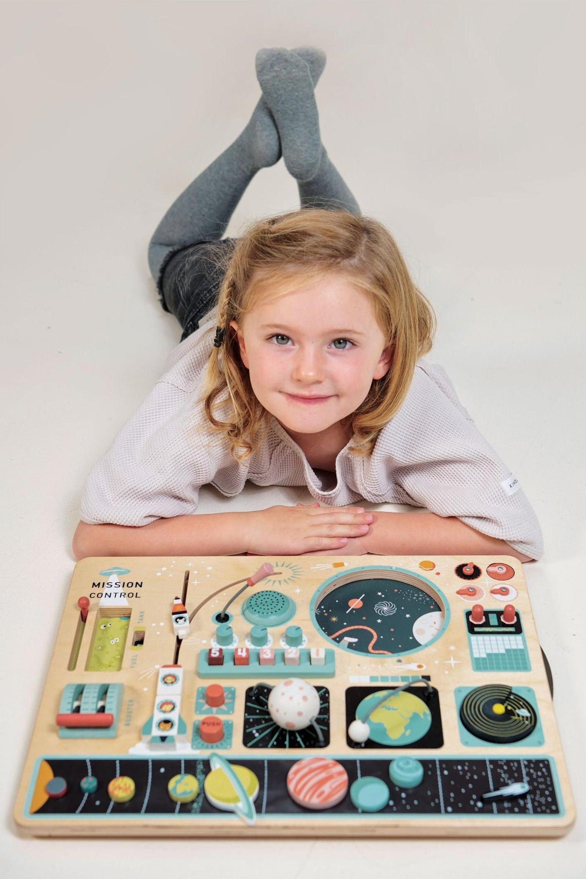 A young child with curly hair lies on the floor, engaging with a colorful wooden space station activity board.