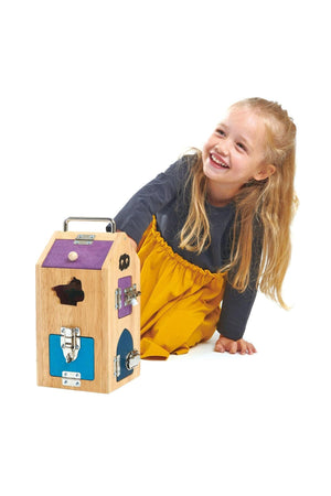 A smiling child in a gray top and yellow skirt interacts with a wooden Monster Lock Box featuring various locking.