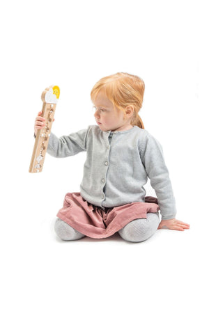 A child in a gray sweater and pink skirt holds a wooden rainmaker toy with cloud and sun design.