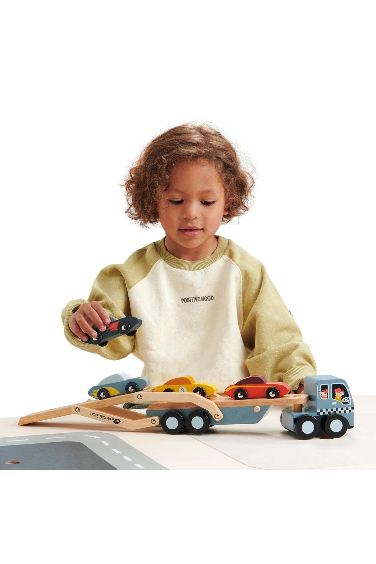A child plays with a Tender Leaf wooden car transporter, featuring colorful super cars and a detachable carrier. The setting emphasizes sustainable wooden construction and safe play features.