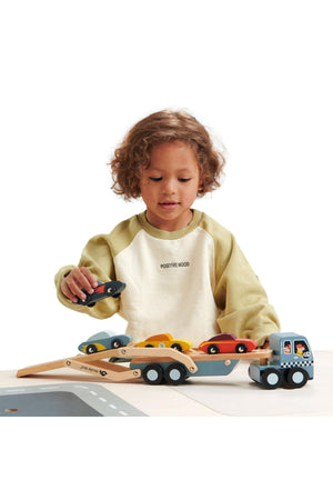 A child plays with a Tender Leaf wooden car transporter, featuring colorful super cars and a detachable carrier. The setting emphasizes sustainable wooden construction and safe play features.