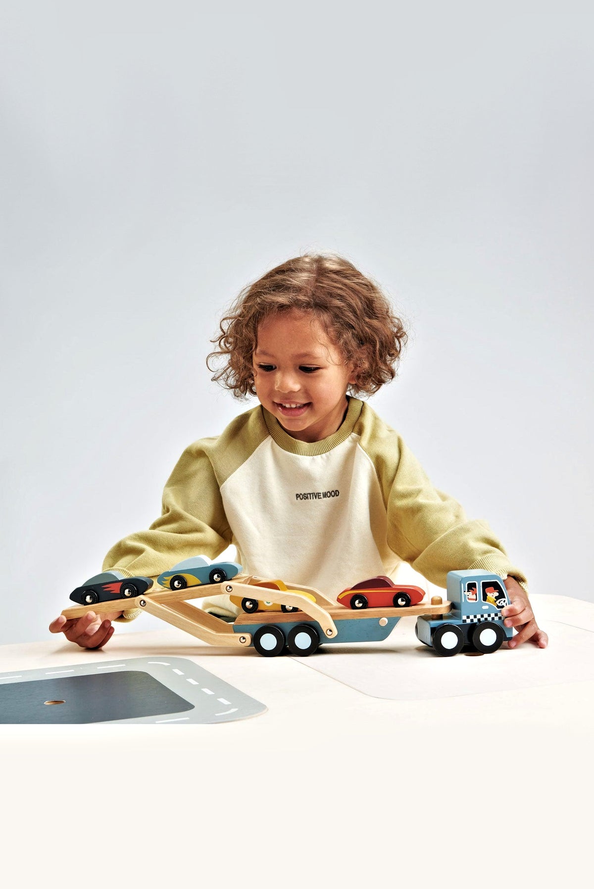 A young child with curly hair plays with a wooden car transporter toy, featuring colorful super cars and a detachable carrier.