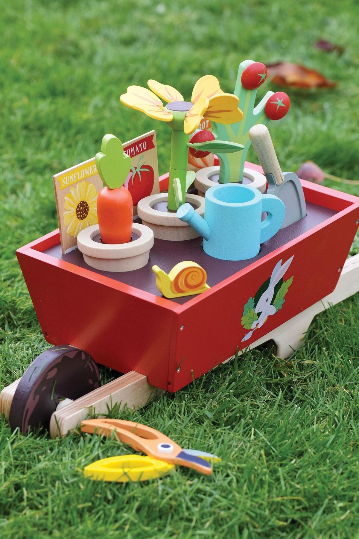 A red wooden wheelbarrow set features flower pots with colorful plants, a watering can, trowel, and gardening tools on grass.