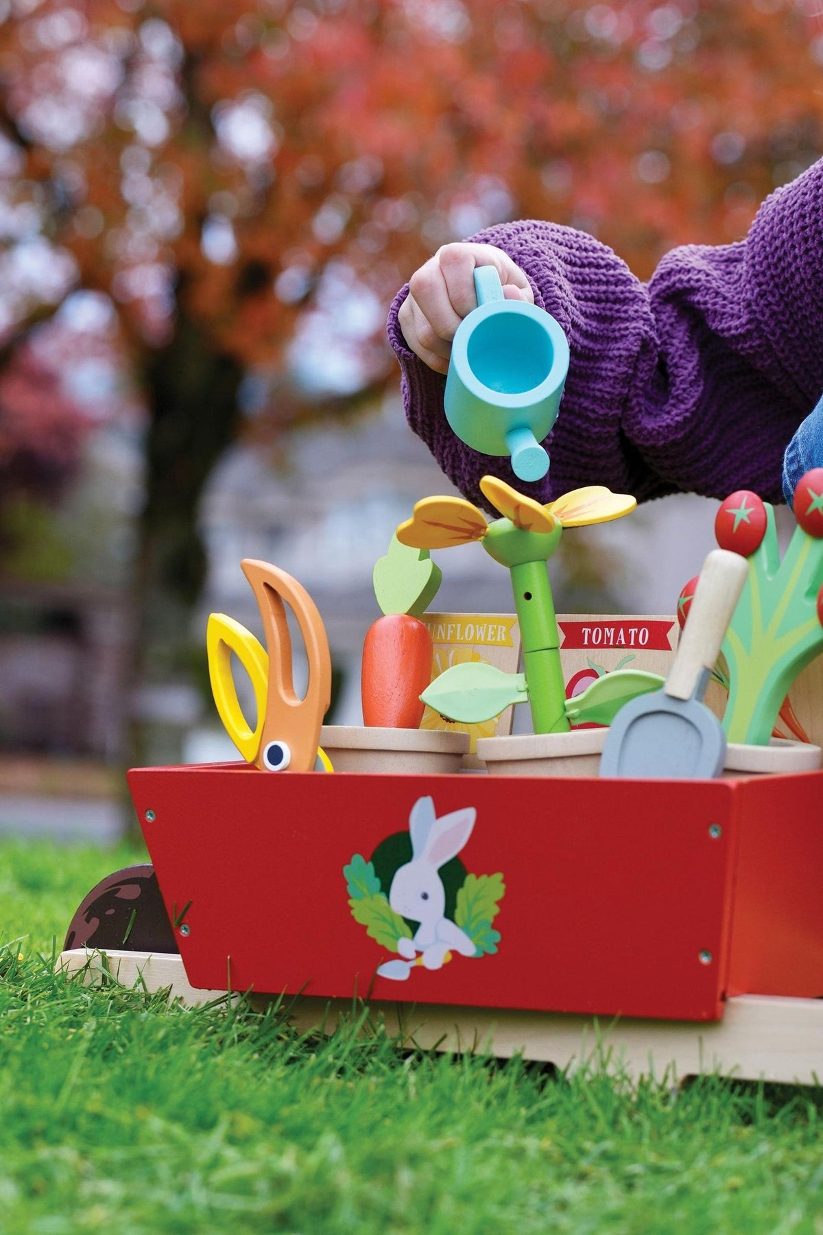 A child waters colorful wooden plants in a red wheelbarrow set, surrounded by green grass and autumn foliage.