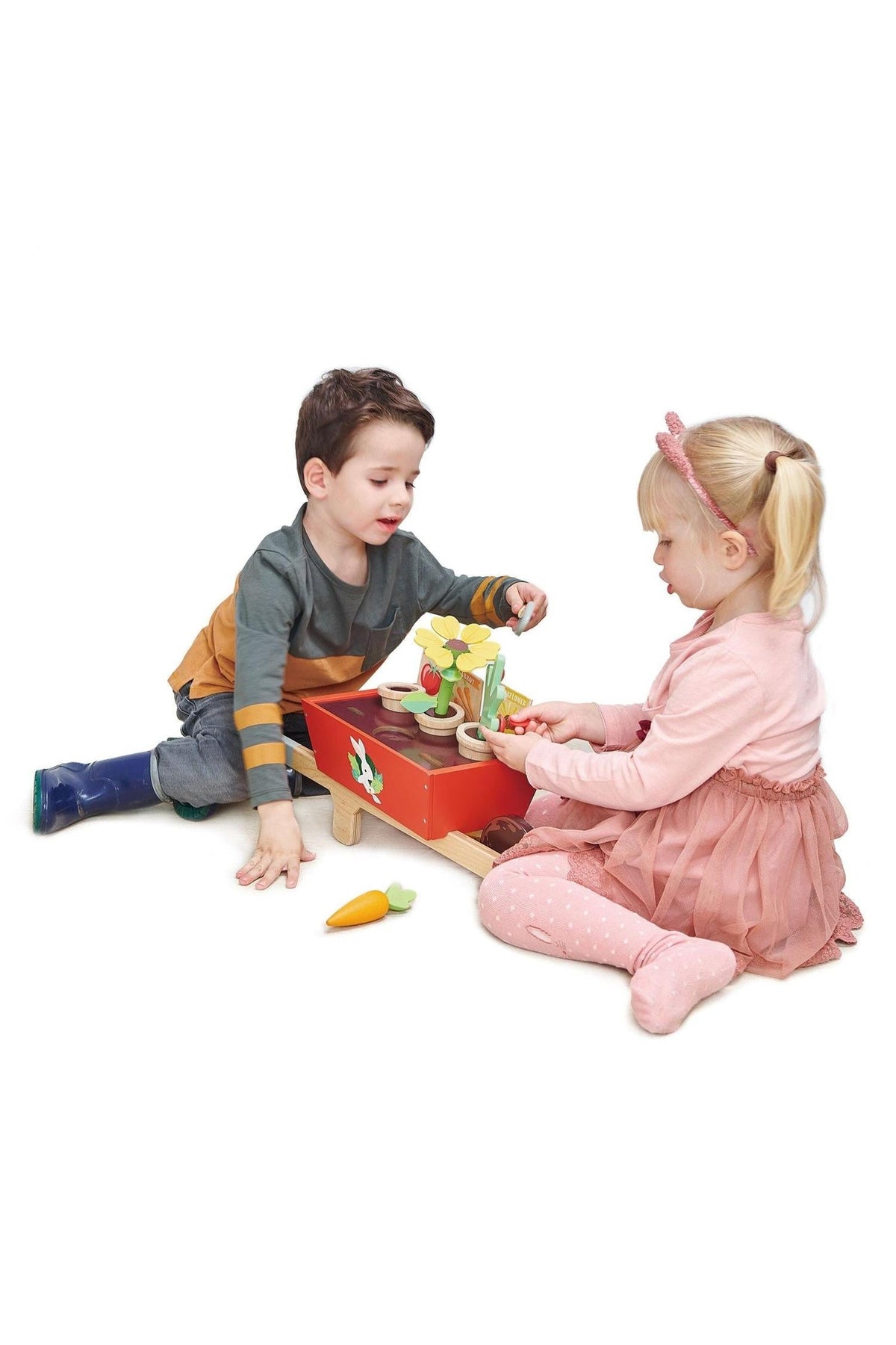 Two children play together with a colorful wooden gardening set, including flower pots and toy vegetables, on a white background.
