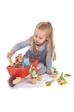 A young girl plays with a red wooden wheelbarrow set, including flower pots, seed packets, and gardening tools.