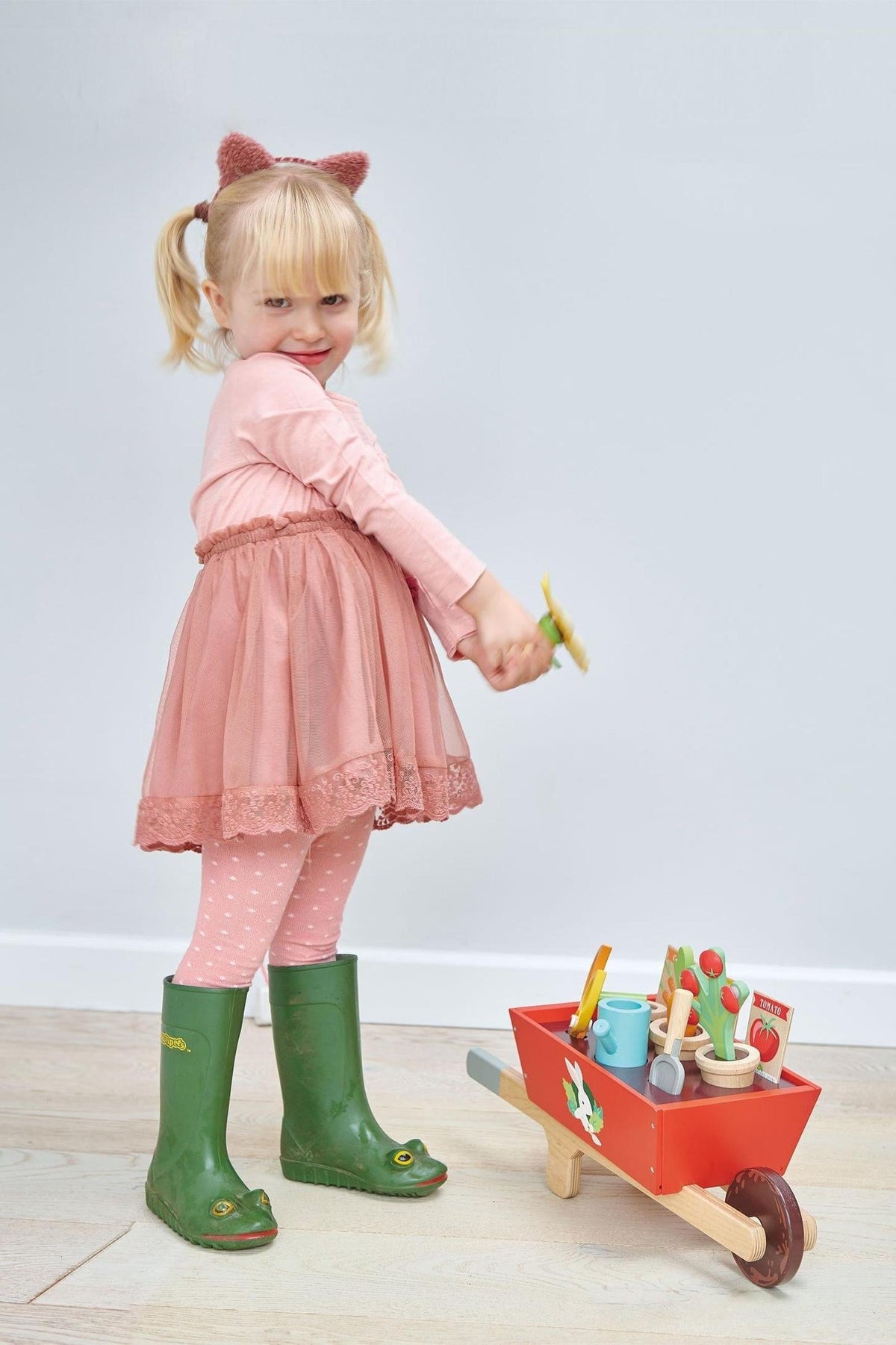 A young girl in a pink dress and green boots holds a toy watering can next to a red wooden.
