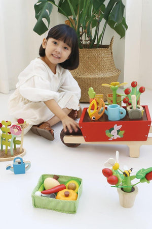 A young child plays with a colorful wooden garden wheelbarrow set, featuring flower pots, gardening tools, and vibrant toy vegetables.